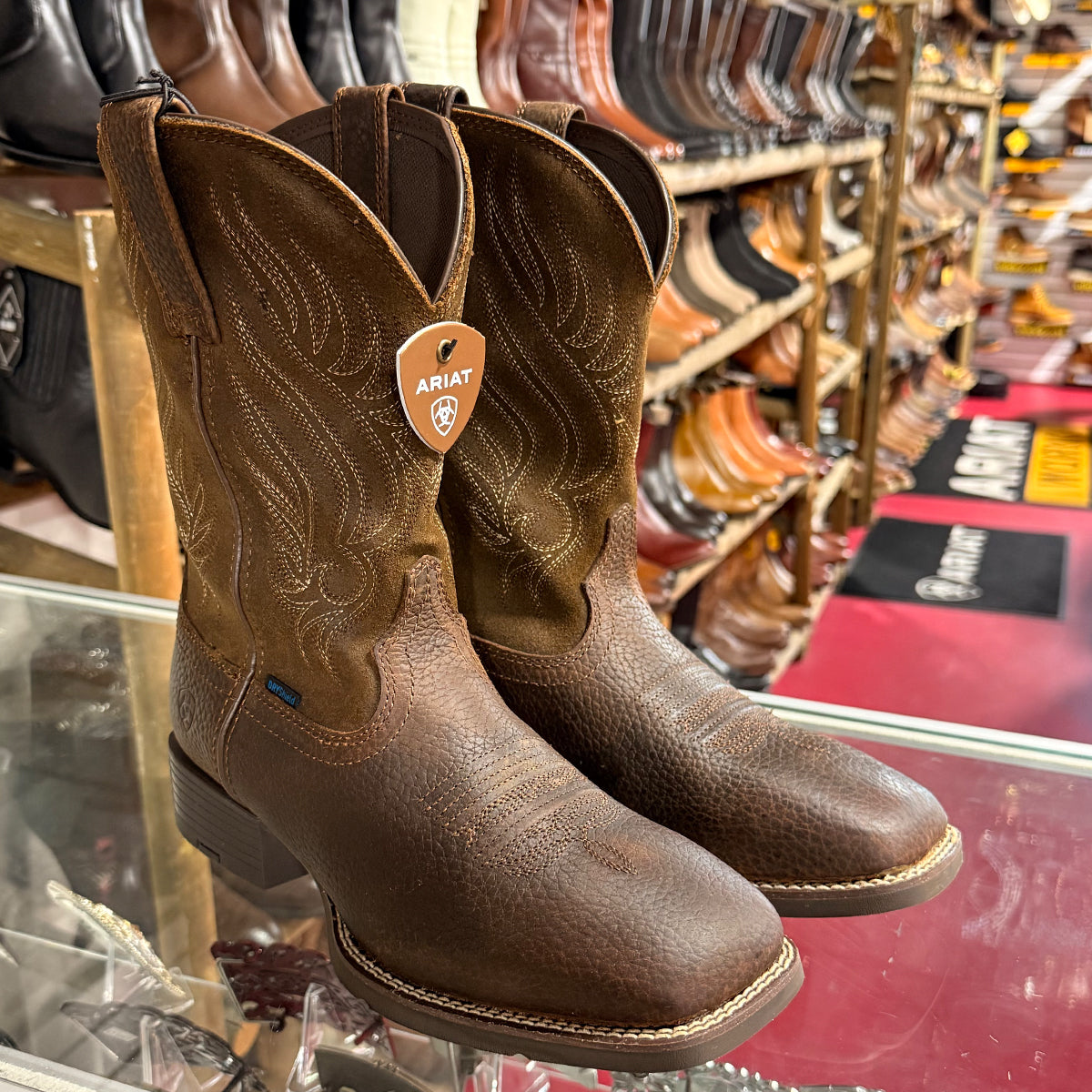 A pair of Ariat brown leather cowboy boots displayed on a glass counter inside a western store, with other boots visible on shelves in the background.