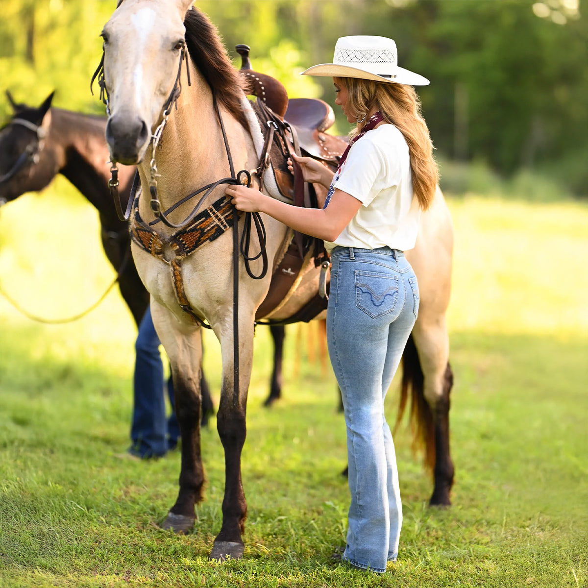 A woman in a white cowboy hat, white t-shirt, and light blue jeans stands on a grassy field next to a light brown horse, adjusting its western saddle and bridle. Another horse is visible in the background.