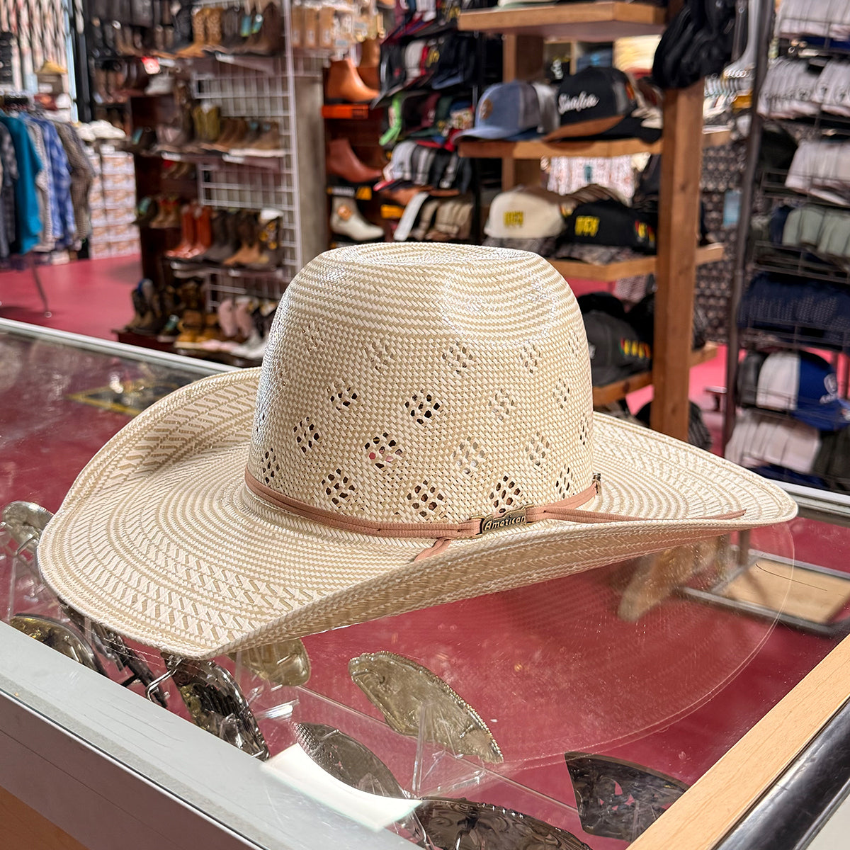 American Hat Company straw western hat with eyelet cutouts and a light pink ribbon band, displayed on a glass jewelry counter inside a retail store.