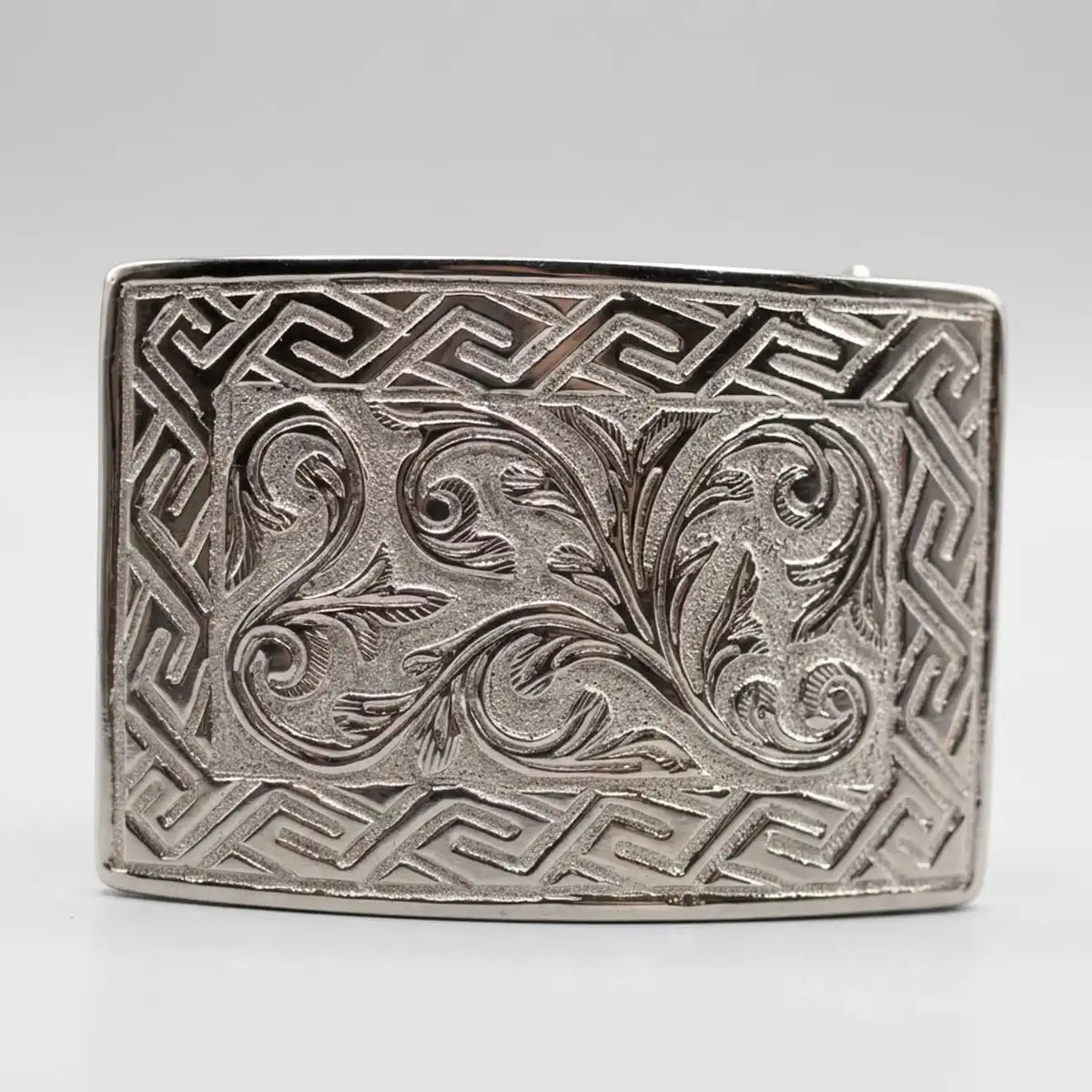 A rectangular silver-toned metal buckle featuring an embossed Greek key border and a central floral scroll motif, shown on a neutral light gray background.
