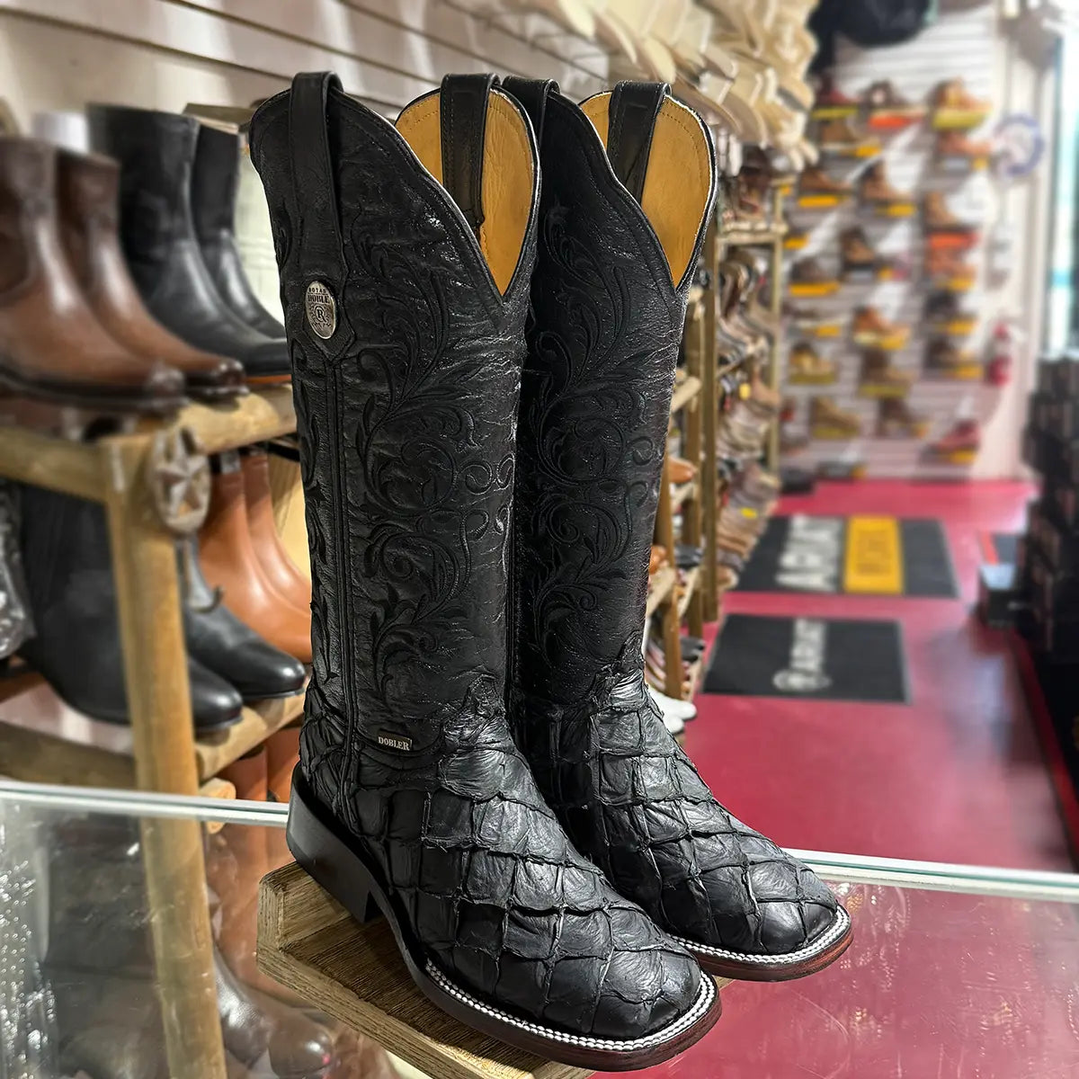 Angled front view of black Doble R pirarucu cowgirl boots displayed on a wooden stand in a western apparel retail store, highlighting the square toe and the tall, intricately embroidered black leather shaft.