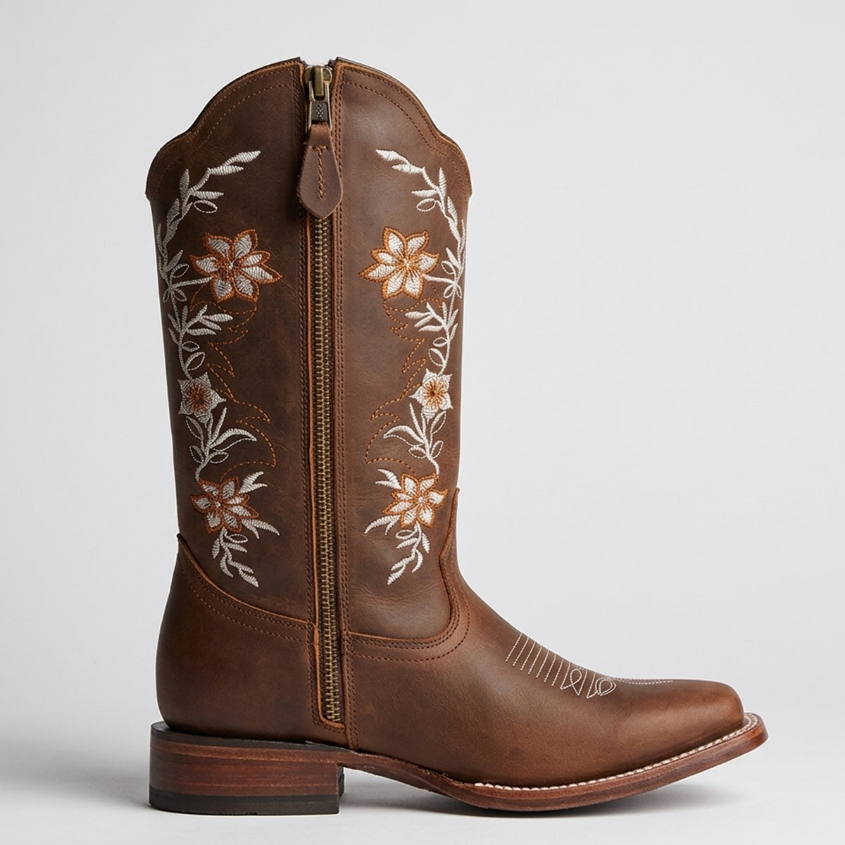 A brown leather cowgirl boot with a side zipper, featuring white and orange floral embroidery on the shaft, set against a light gray background.